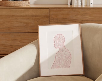 May include: A framed art print featuring a red line drawing of a person's profile. The artwork is set against a white background and framed in a natural wood frame. The print is displayed on a beige armchair.