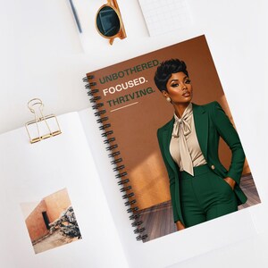 Puede incluir: Cuaderno de espiral con una cubierta marrón que presenta a una mujer con un traje verde. El texto "UNBOTHERED. FOCUSED. THRIVING." está impreso en la cubierta. El cuaderno está abierto, revelando una página en blanco y un clip dorado.