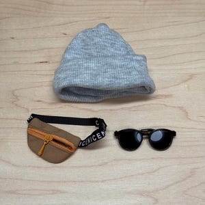 May include: A gray ribbed knit beanie, a brown fanny pack with an orange zipper, and black sunglasses with round frames are displayed on a wooden surface. The fanny pack has a black strap with the word "CENICEN" printed on it.