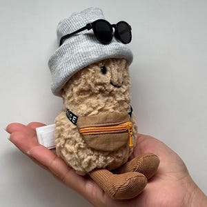 May include: A plush peanut toy with a gray beanie, black sunglasses, and a brown fanny pack. The fanny pack has a gold zipper and the word "NICE" is printed on a tag. The toy has brown corduroy feet and a smiling face.