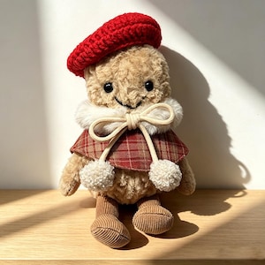May include: A plush doll with tan fuzzy fur, black bead eyes, and a stitched smile. It wears a bright red crocheted beret, a white faux fur collar with a cream-colored drawstring bow, a red plaid capelet, and tan corduroy fabric legs.