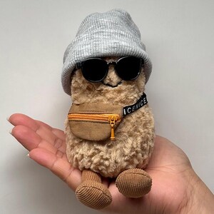 May include: A plush toy shaped like a peanut, wearing a gray beanie, black sunglasses, and a brown fanny pack with the word "ICENICE" on the strap. The toy has corduroy feet and a smiling face.