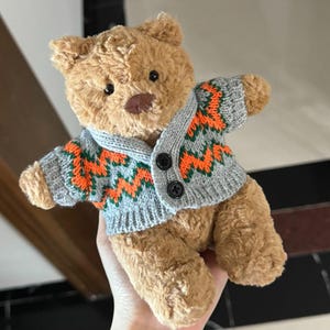May include: A plush teddy bear with brown fur wearing a gray knitted cardigan. The cardigan features an orange and green zigzag pattern and black buttons. The bear has black eyes and a brown nose.