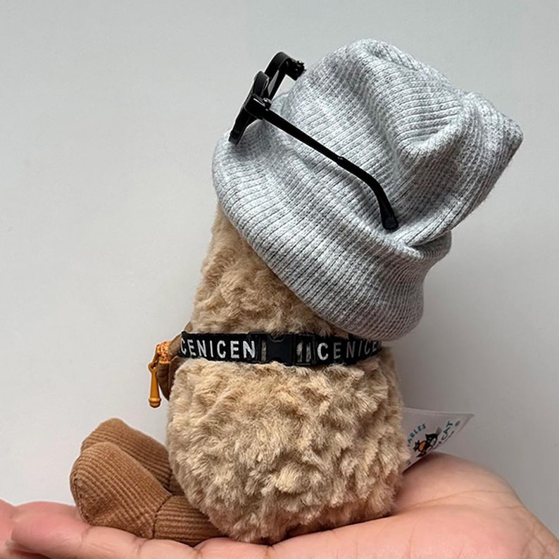 May include: A plush toy with a light brown body, wearing a gray beanie and black-framed glasses. The toy has a black collar with the word "CENICEN" printed on it. The toy is being held in a hand.