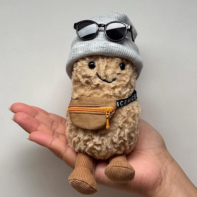 May include: A plush peanut toy with a gray beanie, black sunglasses, and a brown fanny pack. The toy has a smiling face and corduroy feet. The fanny pack has the word "ICENICE" printed on the strap.
