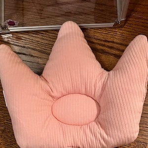 May include: A light pink, crown-shaped baby pillow with a ribbed texture. The pillow has a central, oval-shaped depression and three pointed tips. A clear, glass-like box is in the background.