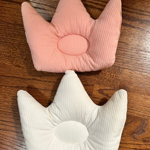 May include: Two crown-shaped baby pillows. One is light pink, the other is white. Both pillows have a ribbed texture and a circular indentation in the center. The pillows are on a wooden surface.