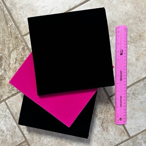 May include: Two square velvet boxes, one black and one hot pink, are stacked at an angle. A pink ruler with white markings and the text "WESTCOTT" is positioned to the right of the boxes. The boxes are on a tiled surface.