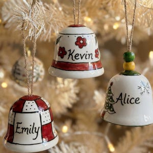 May include: Three ceramic Christmas bell ornaments suspended by twine. Each bell is white with red detailing and personalised names: "Emily", "Kevin", and "Alice". The bells feature floral and Christmas tree designs.