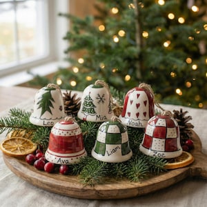 May include: A collection of six ceramic Christmas bells with personalized names, each adorned with unique designs like trees, hearts, and checkered patterns. The bells are arranged on a wooden tray with greenery and cranberries, creating a festive holiday display.