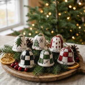 May include: Six ceramic bell ornaments with hand-painted designs including green trees, red hearts, and black, green, or red checkered patterns. Each bell features a name written in black script: Leo, Mia, Noah, Ava, Liam, and Zoe, with twine loops.