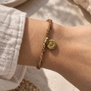 May include: A brown knotted cord bracelet featuring a small, circular, textured brass charm with a sunburst pattern. The charm is attached to the cord with a brass connector, and the bracelet is secured with a series of intricate, hand-tied knots.
