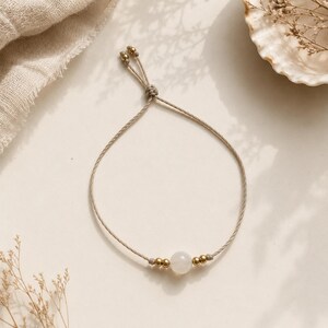 May include: A delicate bracelet featuring a single smooth, translucent white stone bead centred between two small metallic gold-toned beads on each side. The bracelet is constructed from a thin, light beige cord with an adjustable sliding knot closure.