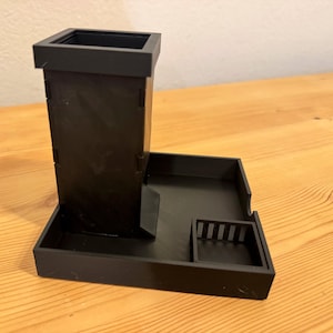 May include: A matte black dice tower featuring a tall, square-shaped vertical chute attached to a rectangular base tray. The base includes a small, square-walled compartment with vertical slats on one side, designed to hold dice after they exit the tower.