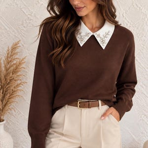 Könnte beinhalten: Ein langärmeliger dunkelbrauner Pullover mit einem weißen spitzen Kragen, der mit klaren blumenförmigen Kristallen verziert ist. Der Pullover wird mit einer beigen Hose und einem dünnen braunen Ledergürtel mit goldener Schnalle kombiniert.