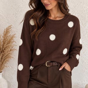 Könnte beinhalten: Ein langärmeliger brauner Pullover mit Rundhalsausschnitt und einem wiederkehrenden Muster aus großen, flauschigen weißen Punkten. Der Pullover wird mit einer dunkelbraunen Hose kombiniert, die einen braunen Ledergürtel mit goldfarbener Metallschnalle hat.
