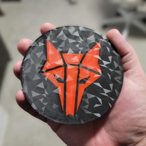 May include: A black and orange 3D printed coaster with a geometric wolf design.