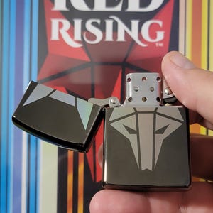May include: A black metal lighter featuring a wolf design and the words "RED RISING" in white. The lighter is open, showing its interior. The background displays a book cover with the same title.