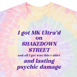 May include: A tie-dye t-shirt in pastel colors with the text "I got MK Ultra'd on SHAKEDOWN STREET and all I got was this t-shirt and lasting psychic damage" in purple. The shirt has a crew neck and short sleeves.