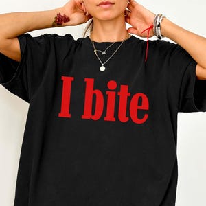 Könnte beinhalten: Ein schwarzes kurzärmeliges T-Shirt mit dem Text "I bite" in fetten roten Buchstaben auf der Brust. Das Kleidungsstück hat einen Rundhalsausschnitt und eine lockere, weit geschnittene Passform, die bis zur Mitte der Oberschenkel reicht.