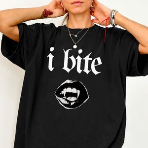 Puede incluir: Una camiseta negra de manga corta con el texto blanco "i bite" en una fuente gótica sobre una ilustración gráfica de un par de labios abiertos con colmillos visibles. La camiseta tiene cuello redondo y un corte holgado y relajado.