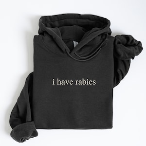 Può includere: Felpa con cappuccio nera con la scritta bianca "i have rabies". La felpa ha un cappuccio con coulisse e maniche lunghe. La parola "BLACK" è visibile in basso a destra.