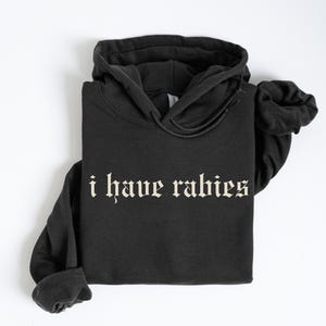 Puede incluir: Sudadera con capucha negra con el texto "i have rabies" impreso en una fuente gótica blanca.