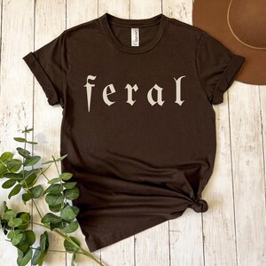 以下が含まれることがあります： 「feral」の文字がオフホワイトのスタイリッシュなフォントでプリントされた、濃い茶色のTシャツ。Tシャツは半袖でクルーネック。Tシャツは裾で結ばれています。背景には茶色の帽子と緑の葉があります。