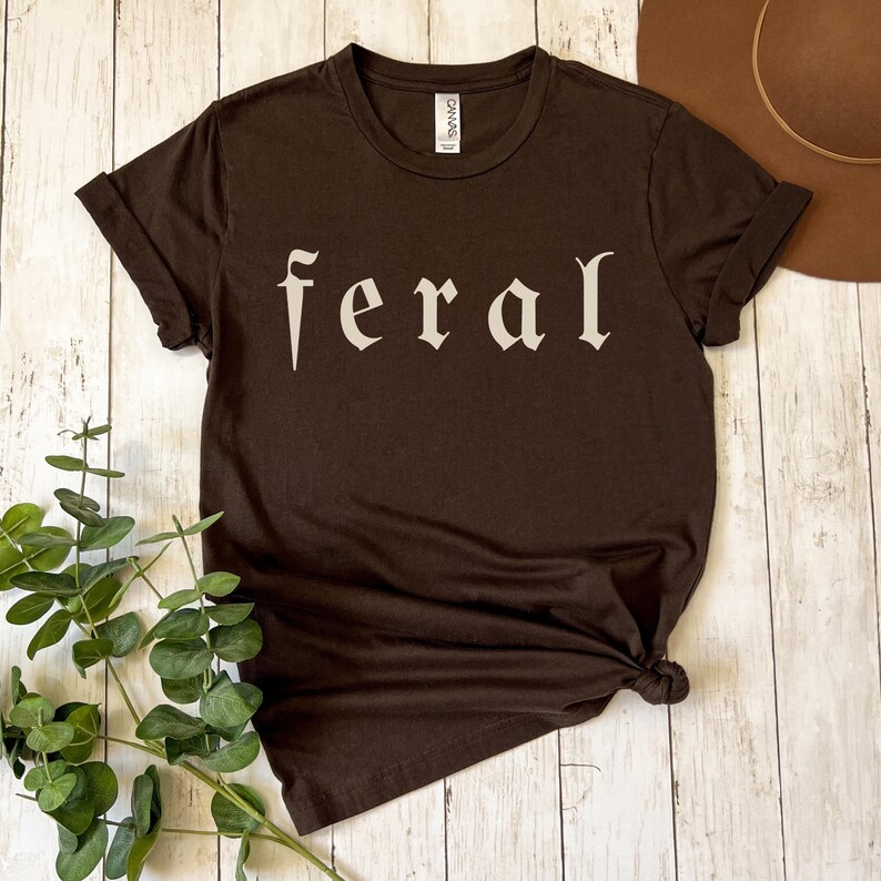 以下が含まれることがあります： 「feral」の文字がオフホワイトのスタイリッシュなフォントでプリントされた、濃い茶色のTシャツ。Tシャツは半袖でクルーネック。Tシャツは裾で結ばれています。背景には茶色の帽子と緑の葉があります。