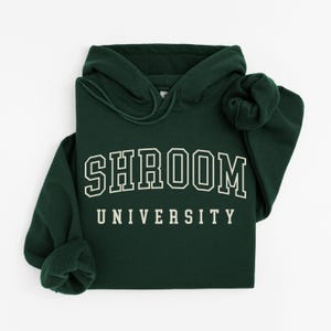 May include: A dark green hoodie with the text "SHROOM UNIVERSITY" in white block letters. The hoodie has a drawstring hood and long sleeves. The garment is folded neatly on a white surface.