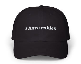 May include: Black baseball cap with the embroidered text "i have rabies" in white. The cap has a curved brim and a six-panel design. The text is in a cursive font.