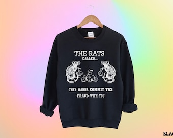 May include: Black sweatshirt with the text "THE RATS CALLED... THEY WANNA COMMIT TAX FRAUD WITH YOU." The design features two rats holding money bags and a pile of money bags. The sweatshirt is hanging on a wooden hanger.