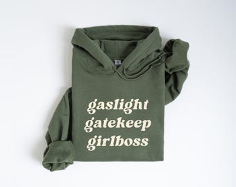 Puede incluir: Una sudadera con capucha verde con el texto "gaslight gatekeep girlboss" impreso en color crema.