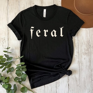 以下が含まれることがあります： ゴシック体で「feral」の文字がプリントされた黒いTシャツ。袖は折り返されています。Tシャツは裾で結ばれています。Tシャツは柔らかい素材で作られています。