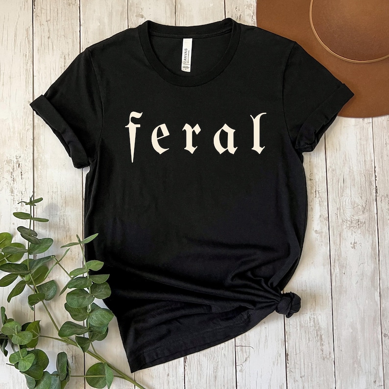 以下が含まれることがあります： ゴシック体で「feral」の文字がプリントされた黒いTシャツ。袖は折り返されています。Tシャツは裾で結ばれています。Tシャツは柔らかい素材で作られています。