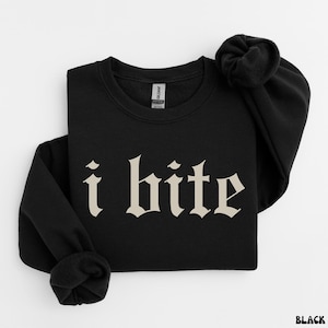 May include: Black sweatshirt with the phrase "i bite" in a gothic font. The text is in a light beige color, contrasting with the dark fabric. The sweatshirt is folded, with the sleeves and collar visible.