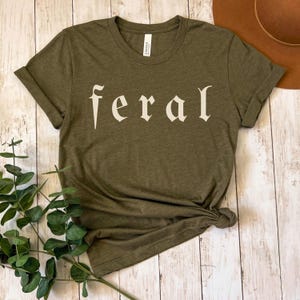 以下が含まれることがあります： オリーブグリーンのTシャツ。オフホワイトのスタイリッシュなフォントで「feral」の文字がプリントされています。裾は結ばれ、袖は折り返されています。背景には茶色の帽子が見えます。