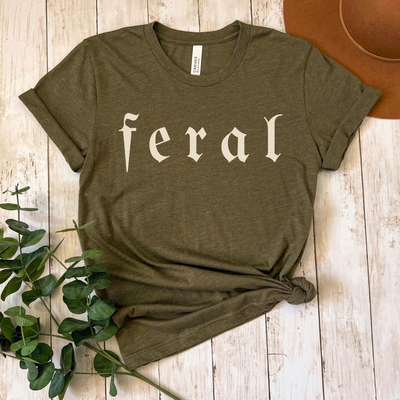 以下が含まれることがあります： オリーブグリーンのTシャツ。オフホワイトのスタイリッシュなフォントで「feral」の文字がプリントされています。裾は結ばれ、袖は折り返されています。背景には茶色の帽子が見えます。