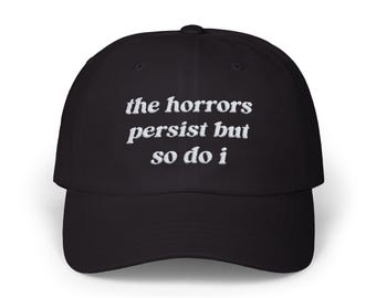 May include: Black baseball cap with white embroidered text that reads "the horrors persist but so do i"