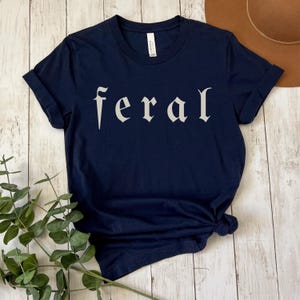 以下が含まれることがあります： ゴシック体で「feral」の文字がプリントされたネイビーブルーのTシャツ。 シャツは裾で結ばれており、袖は折り返されています。 背景には木製の表面と茶色の帽子が含まれています。