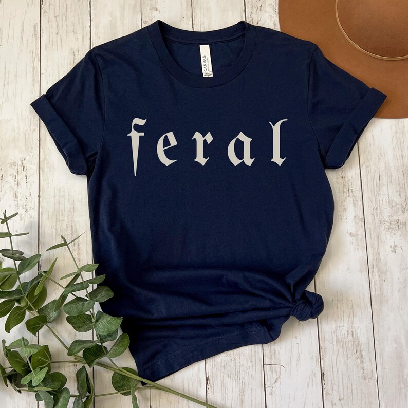 以下が含まれることがあります： ゴシック体で「feral」の文字がプリントされたネイビーブルーのTシャツ。 シャツは裾で結ばれており、袖は折り返されています。 背景には木製の表面と茶色の帽子が含まれています。