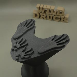 Könnte beinhalten: Ein schwarzer 3D-gedruckter Stempel mit erhabenen Tierpfotenabdrücken auf der Oberfläche. Der Stempel hat einen breiten, texturierten oberen Teil und einen zylindrischen Sockel. Im Hintergrund ist der Text WILD DRUCK in beigen 3D-Buchstaben zu sehen.