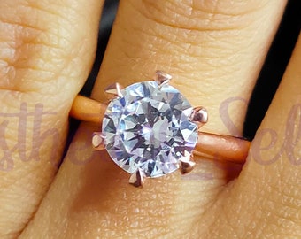 May include: A rose gold engagement ring with a large, clear, round-cut center stone. The ring is on a finger.