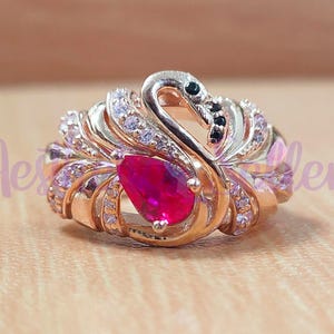May include: Rose gold swan ring featuring a pear-shaped, deep red gemstone. The swan's head is black and the body is adorned with small, clear gemstones. The ring has a detailed, ornate design.