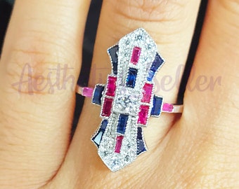 Könnte beinhalten: Ein silberner Ring mit geometrischem Design, der einen großen Mittel-Stein und kleinere Steine in einem geometrischen Muster zeigt. Der Ring ist mit blauen und roten Edelsteinen verziert.
