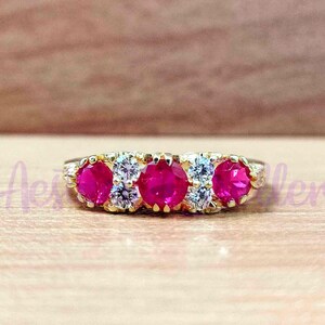 May include: A gold ring featuring five vibrant red gemstones, interspersed with smaller, clear stones. The ring is set against a warm, wooden background, showcasing the jewelry's design and color.