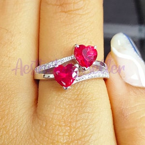 May include: A silver ring with two red heart-shaped gemstones. The ring has a delicate design with small clear stones.