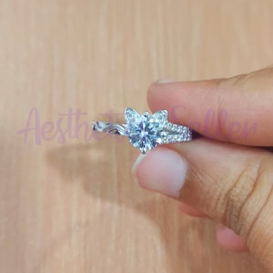 May include: A silver ring with a heart-shaped, clear gemstone centre. The ring features a split band with smaller, clear stones and a floral design around the main stone. The word "Aesthetic Seller" is visible in the background.