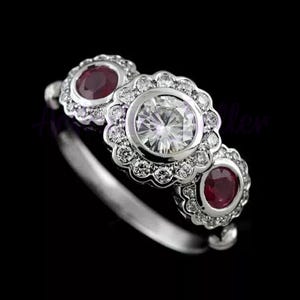 May include: A silver ring with three floral-shaped settings. The center setting holds a large, clear gemstone, flanked by two smaller settings with red gemstones. The settings are surrounded by small, clear stones.