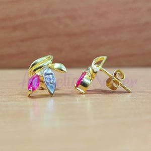 Könnte beinhalten: Ein Paar goldfarbene Ohrstecker. Ein Ohrring hat ein Blattdesign mit einem rosa Edelstein und einem kleinen, klaren Stein. Der andere Ohrring hat einen einzelnen, rosa Edelstein. Die Ohrringe liegen auf einer Holzoberfläche.
